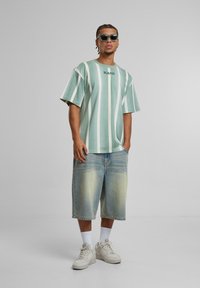 Grønn og hvit stripete oversized t-skjorte med "KANI" logo, kombinert med lyseblå denimshorts og hvite joggesko. Teksturert stoff, uformell design.