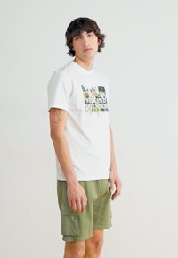 Weißes kurzärmliges T-Shirt mit einem grafischen Druck, der "WEEKEND OFFENDERS" über einem mehrfarbigen Design zeigt; kombiniert mit olivgrünen Cargo-Shorts.