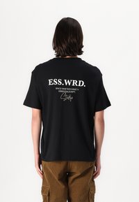 Fekete pamut póló rövid ujjakkal, fehér felirattal a hátán: "ESS.WRD. SINCE NINETEEN NINETY ESSENTIALS DEPT. Studio."