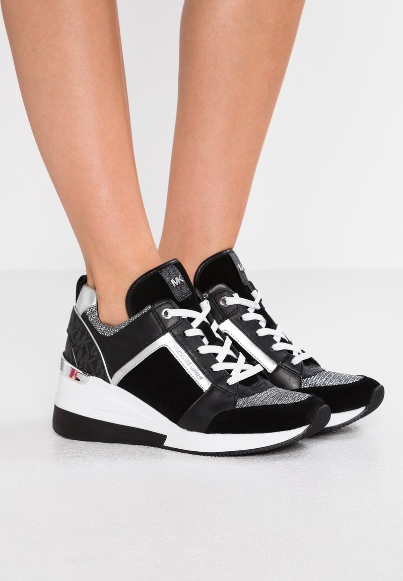 Des pieds portant des baskets Michael Kors noires et argentées avec des lacets blancs sur un sol gris clair contre un fond blanc.