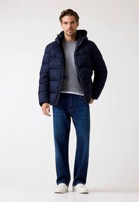 Marineblaue Steppjacke mit Frontreißverschluss, Kapuze und gesteppter Textur; kombiniert mit grauem T-Shirt und Jeans. Weiße Sneakers vervollständigen den Look.