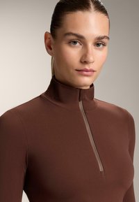 Cămașă cu gât înalt și mâneci lungi, realizată dintr-un material neted, de culoare maro, cu un fermoar pe jumătate ce are un accent bej de-a lungul fermoarului. Design simplu.