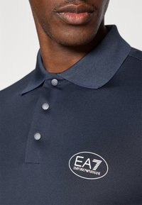 Jūras zils polo krekls, kas izgatavots no elpojoša auduma, ar divu pogu aizdari, īsām piedurknēm un izšūtu EA7 logotipu uz krūšu daļas.