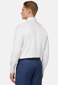 Chemise blanche à manches longues avec un col évasé, texture lisse et accents de poignets boutonnés, associée à un pantalon bleu ajusté.