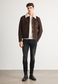 Hnedá semišová bunda s krémovým umelým kožúškom na golieri, dvoma náprsníkovými vreckami, spárovaná s krémovým svetrom a čiernymi slim-fit džínsami. Čierne členkové čižmy.