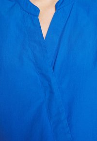 Blaues Oberteil mit V-Ausschnitt aus glattem Stoff. Verfügt über präzise Nähte und eine schlichte, klare Silhouette.