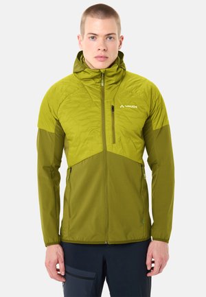 Giovane uomo che indossa una giacca outdoor Vaude con cappuccio in due tonalità di verde, in piedi frontalmente con le braccia rilassate lungo i fianchi.