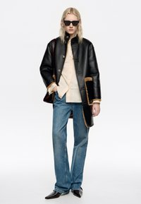 Persona vistiendo un abrigo de cuero negro con ribete de piel de tono marrón claro, camisa crema, jeans azules, zapatos negros de punta, gafas de sol negras y llevando un bolso marrón oscuro.