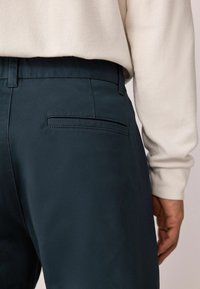 Pantalons bleu sarcelle foncé avec une texture lisse, dotés d'une poche arrière unique et d'une taille ajustée, associés à un haut léger à texture.
