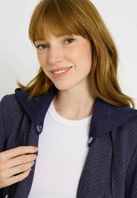 Chaqueta azul marino con tela texturizada y capucha en contraste. Camiseta blanca de canalé debajo. La chaqueta tiene detalles de botones.