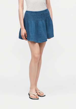 Žena nosi modrú textúrovanú minisukňu so záhybovaným pásom a béžové žabky, stojí na jednoduchom pozadí.