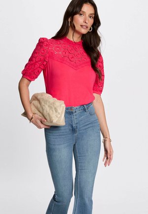 Top rosa brillante con maniche corte ricamate in pizzo, abbinato a jeans a campana azzurri. Tiene in mano una borsa a tracolla in oro testurizzato.