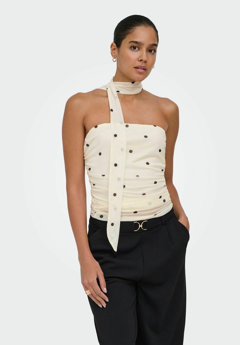 Femme portant un haut blanc sans manches à pois avec une cravate autour du cou et un pantalon noir avec une ceinture à accents dorés, posant avec une main dans la poche.