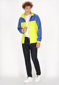 Chaqueta cortavientos amarilla brillante y azul con cierre frontal, puños elásticos y cuello alzado, combinada con jeans oscuros y zapatillas blancas.