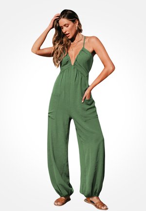Frau trägt einen grünen ärmellosen Jumpsuit mit tiefem V-Ausschnitt und elastischen Bündchen, posiert mit einer Hand in der Tasche und der anderen, die ihr Haar berührt.