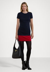 Vestido azul-marinho com mangas curtas, barra vermelha e detalhes em botão dourado nos ombros. Mala preta e botas pretas até ao joelho.