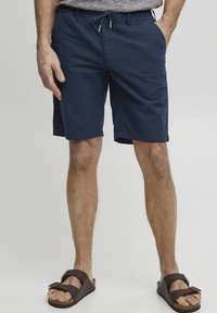Marineblå shorts laget av lett materiale, med elastisk midjebånd og snor, sidelommer og en avslappet passform. Brune sandaler med to stropper.