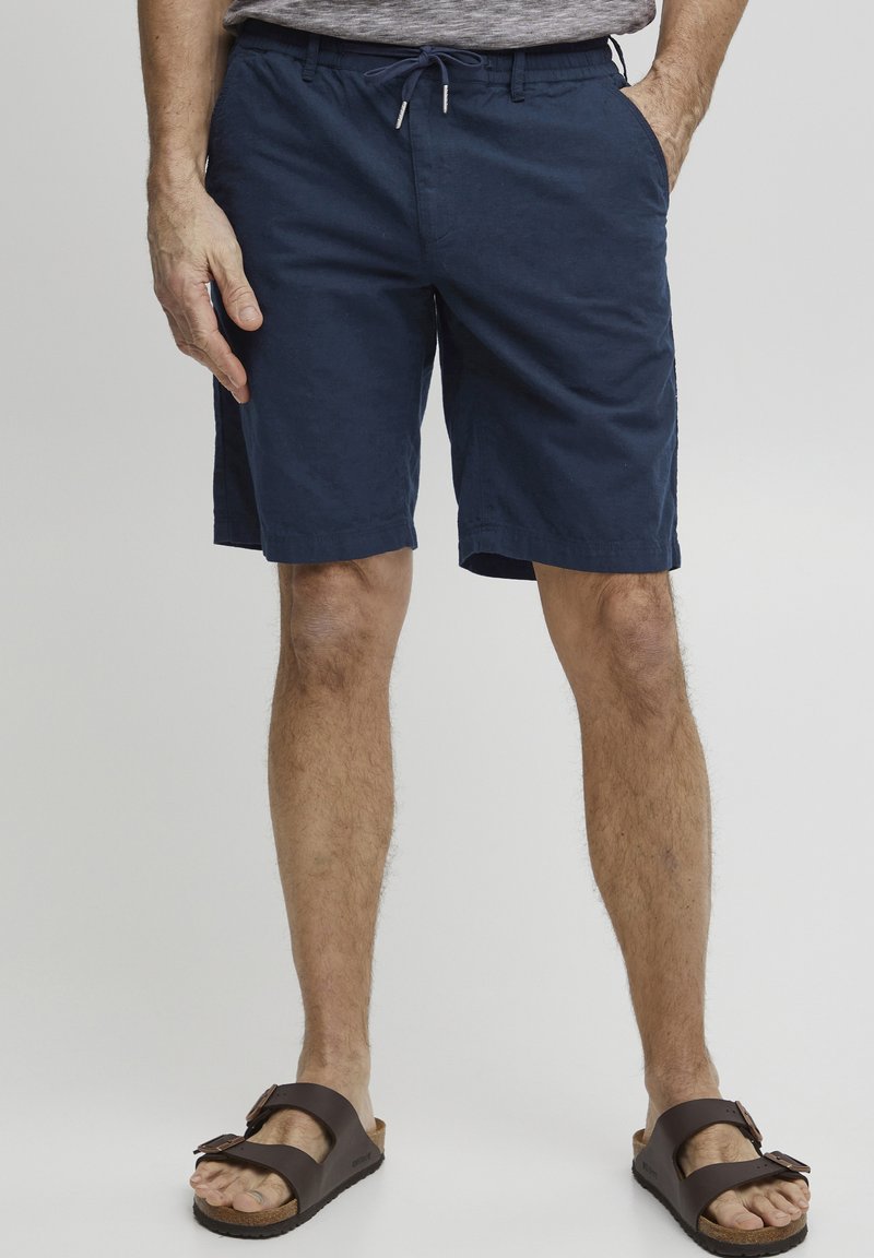 Marineblå shorts laget av lett materiale, med elastisk midjebånd og snor, sidelommer og en avslappet passform. Brune sandaler med to stropper.