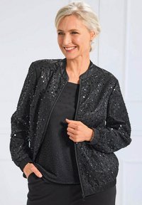 Veste bomber noire ornée de sequins et d'une texture lisse, avec des poignets et un ourlet côtelés, associée à un haut noir.