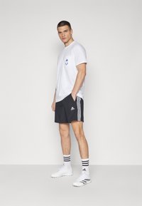 Ung mann står med hendene i lommene iført hvit Adidas t-skjorte, svarte Adidas shorts, hvite Adidas sokker og hvite sportssko.