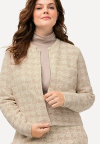 Beige jas met textuur en een subtiel patroon, voorzien van een ritssluiting aan de voorkant en lange mouwen. Draagt over een lichtbeige coltrui, wat zorgt voor een gelaagd uiterlijk.