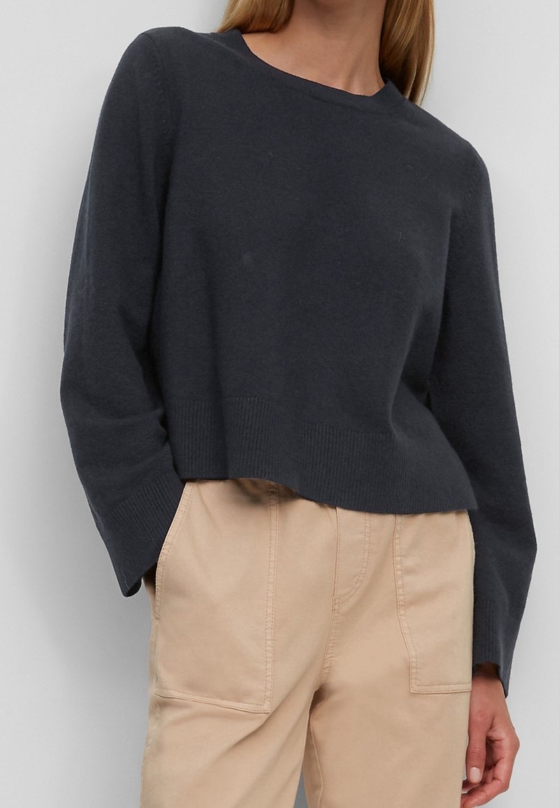 Femme portant un pull gris foncé à manches longues et col rond, rentré dans un pantalon beige taille haute avec de grandes poches avant.