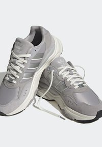 Ett par grå Adidas-sneakers med vita snörningar, tre ränder på sidorna, tjocka vita sulor och Adidas-logotyp på tungan, på vit bakgrund.