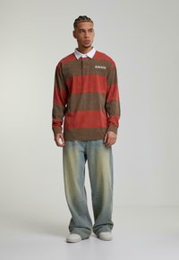 Stripete langermet polo i rød og brun med hvit krage, kombinert med falmede lyseblå jeans og hvite joggesko.