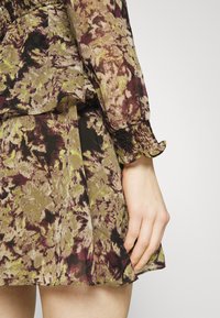 Blumenkleid mit einem Muster in Grün, Lila und Schwarz, ausgestattet mit einem smockierten Bund und Rüschenschäften, aus einem leichten, strukturierten Stoff gefertigt.