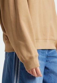 Felpa di cotone beige con polsini a coste, abbinata a jeans blu. Presenta una vestibilità comoda e cuciture visibili lungo l'orlo.