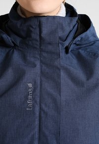Gros plan sur une personne portant une veste d'extérieur Lafuma bleue avec un col haut et un marquage visible sur le devant.