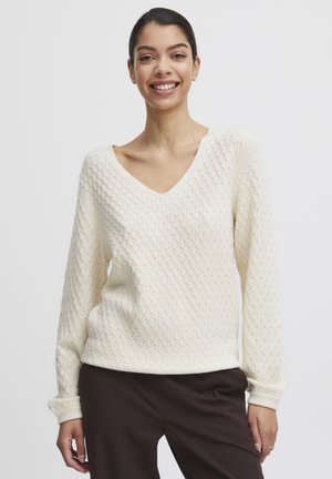 Maglione lavorato a maglia color crema con scollatura a V, maniche lunghe, motivo testurizzato e polsini a costine. Indossato sopra pantaloni marrone scuro.