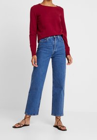 Roter Pullover mit langen Ärmeln kombiniert mit hoch taillierten blauen Jeans mit geradem Bein. Modell trägt schwarze Riemchensandalen. Einfaches, lässiges Outfit.