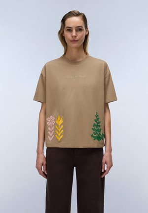 Femme portant un t-shirt beige avec une broderie florale et le texte "Unveiled in Nature", debout devant un fond uni et clair.
