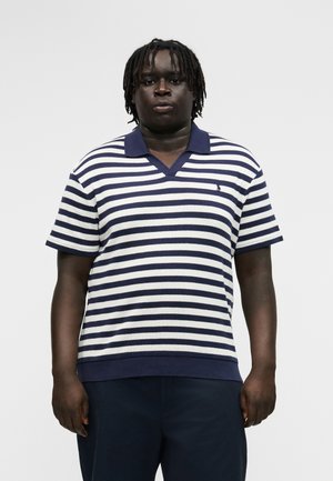 Άνδρας με κοντά dreadlocks, φορώντας κοντομάνικο πόλο μπλουζάκι με οριζόντιες μπλε και λευκές ρίγες και σκούρο παντελόνι, μπροστά σε απλό φόντο.