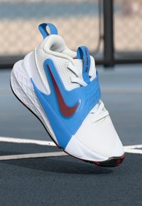Balti krepšinio bateliai su mėlynomis detalėmis, tekstūruota viršutinė dalis, ryškus raudonas Nike logotipas ir išskirtinis geometrinis raštas ant pado.