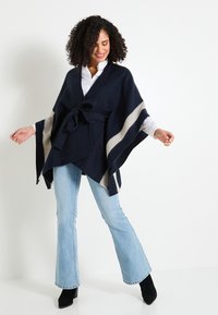 Poncho azul marino hecho de material suave, con un cinturón atado, rayas crema y un diseño de frente abierto. Jeans acampanados y botas de tacón completan el conjunto.