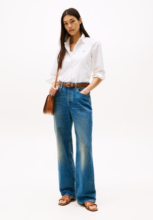Vit långärmad skjorta, blå vida jeans, brunt läderbälte, brun crossbody-väska och bruna sandaler. Slät textur, minimal design.
