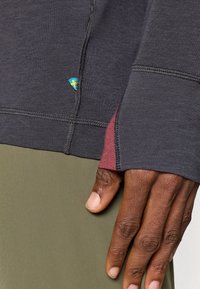 Hosszú ujjú, szürke teljesítmény póló bordó részletekkel; lapos varrásokkal és egy kis logóval rendelkezik. Olíva zöld nadrággal viselve, kéz látható.