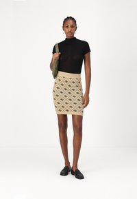 Haut à col roulé noir ajusté associé à une jupe en tricot beige avec un motif en diamant noir. Le mannequin tient un sac vert et porte des mocassins noirs.