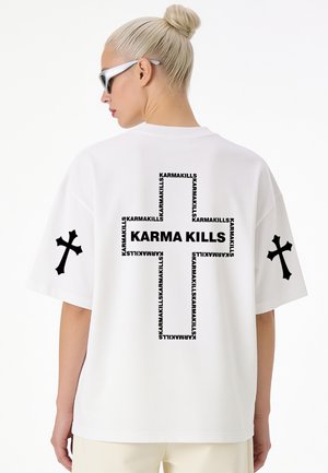 Femme aux cheveux blonds en chignon porte un t-shirt blanc avec une croix noire et le texte "KARMA KILLS" dans le dos ainsi que des croix noires sur les manches.