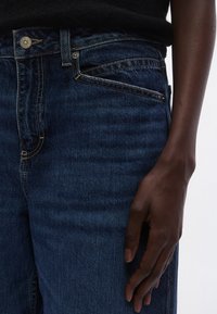 Sötétkék farmernadrág klasszikus kialakítással, kontrasztos varrással, gombolós záródással és elülső zsebbel. Simaságú textúra és testhez álló fazon.