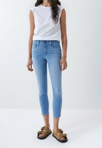 Jeans ajustados de color azul claro con una cintura alta, combinados con una blusa blanca sin mangas ceñida y sandalias marrones con un detalle de lazo.