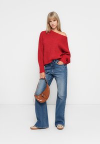 Pull en tricot rouge avec un décolleté asymétrique, associé à un jean large bleu clair. Elle tient un sac à main en cuir texturé marron. Chaussures plates beiges.