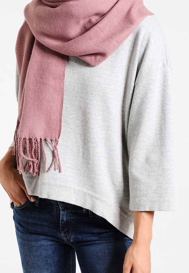 Personne portant un haut à manches longues gris clair, un jean bleu foncé et une écharpe rose à franges drapée autour du cou.