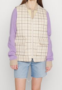 Beige vadderad väst med brunt rutmönster, med två framfickor, bärs över en lila långärmad tröja och denimshorts.