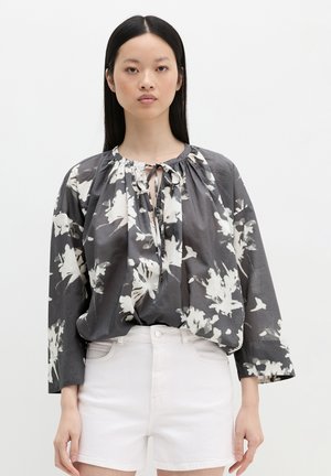 Kobieta ubrana w luźną szarą bluzkę z białymi kwiatowymi wzorami i białe szorty z wysokim stanem na jednolitym tle.