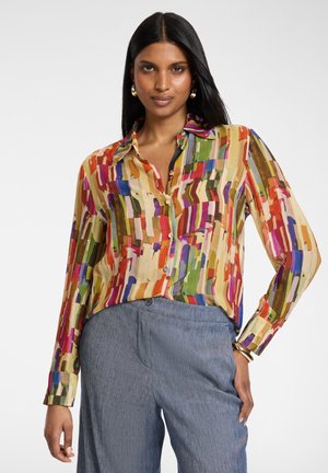 Frau trägt eine farbenfrohe, abstrakt gemusterte Bluse mit Knöpfen und eine blaue, hoch taillierte Hose, steht mit einer Hand in der Tasche.