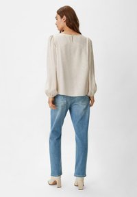 Hellbeige, strukturierte Bluse mit Puffärmeln und einer Knopfleiste hinten, kombiniert mit hellblauen, geraden Jeans und beigen Absätzen.