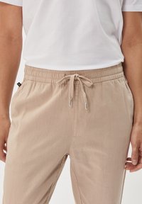 Beige Jogginghose mit elastischem Bund, weichem Stoff, Seitentaschen und silbernen Spitzen an der Kordel. Getragen mit einem weißen T-Shirt.